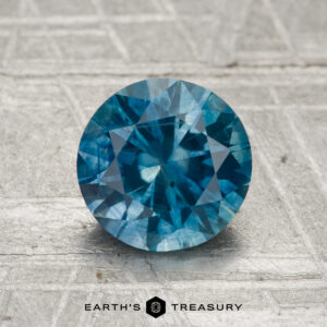 A 1.67-carat heated Montana sapphire with a round cut is displayed on a textured gray surface, featuring the "Earth's Treasury" logo at the bottom.
