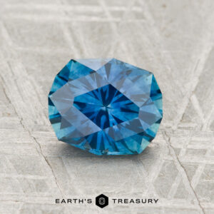A 1.65-carat heated Montana sapphire with a faceted blue hue rests on a textured surface, with the "EARTH'S TREASURY" logo at the bottom.
