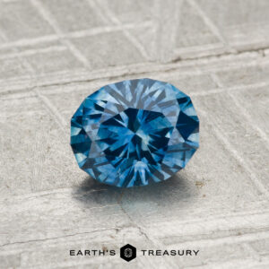 A 1.03-carat heated Montana sapphire rests on a textured metallic surface with "Earth's Treasury" at the bottom.