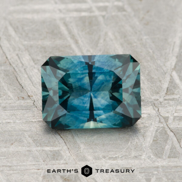 1.67-Carat Montana Sapphire (Heated) 1.67-carat step-cut blue-green Montana sapphire (heated) displayed on textured gray surface; "Earth's Treasury" logo at the bottom.