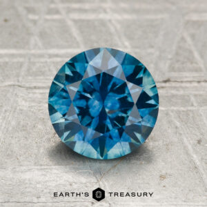 A 1.61-carat heated Montana sapphire, round brilliant-cut and blue in color, displayed on a textured gray surface with the "Earth's Treasury" logo and text below.