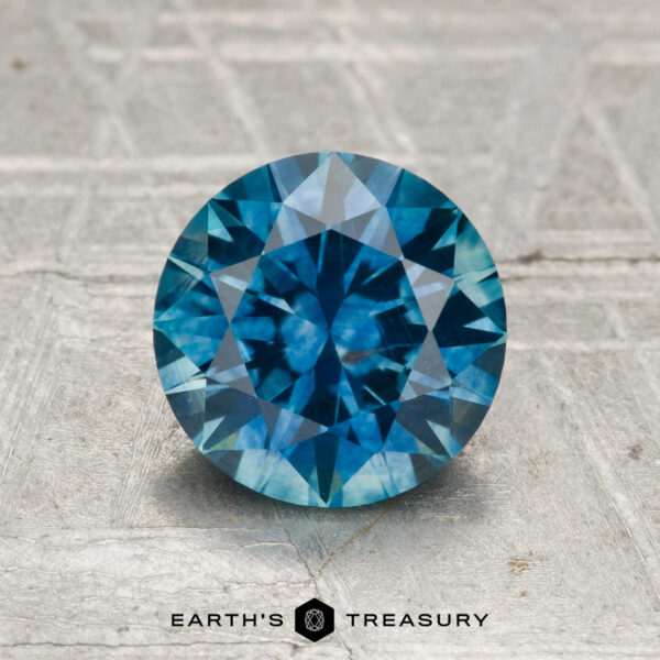 1.61-Carat Montana Sapphire (Heated) A 1.61-carat heated Montana sapphire, round brilliant-cut and blue in color, displayed on a textured gray surface with the "Earth's Treasury" logo and text below.