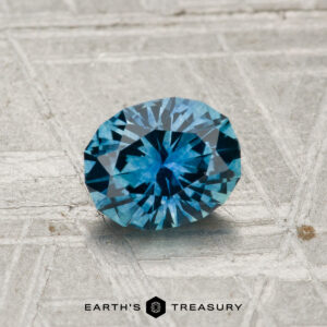 A 1.02-carat heated Montana sapphire with a blue hue is displayed on a textured, light-colored surface above the "Earth's Treasury" logo.