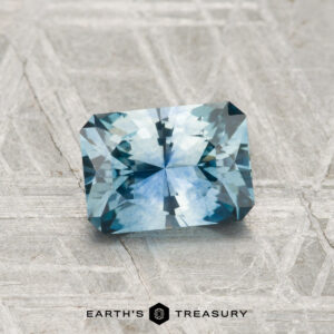 A 1.35-carat blue-green faceted Montana sapphire (heated) is showcased on a textured gray surface, with "EARTH'S TREASURY" text beneath it.