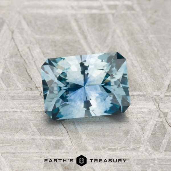 1.35-Carat Montana Sapphire (Heated) A 1.35-carat blue-green faceted Montana sapphire (heated) is showcased on a textured gray surface, with "EARTH'S TREASURY" text beneath it.
