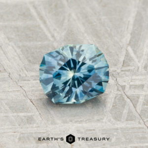 A 1.04-carat heated Montana Sapphire with a blue-green hue is displayed on a textured, light gray surface. "EARTH'S TREASURY" appears at the bottom of the image.