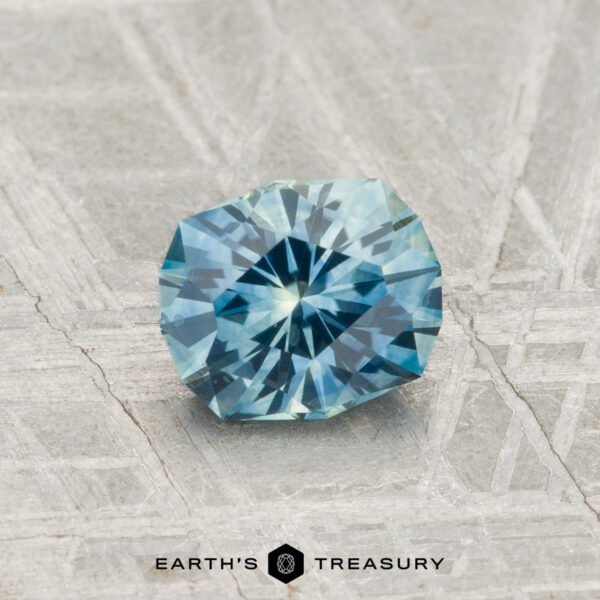 1.04-Carat Montana Sapphire (Heated) A 1.04-carat heated Montana Sapphire with a blue-green hue is displayed on a textured, light gray surface. "EARTH'S TREASURY" appears at the bottom of the image.
