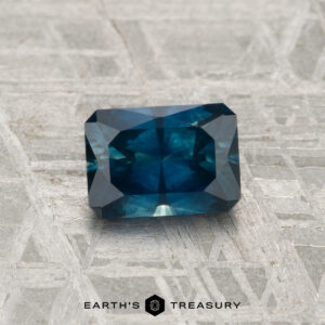 A 0.96-carat heated Montana sapphire with faceted edges is displayed on a textured gray surface, with "EARTH'S TREASURY" text at the bottom.