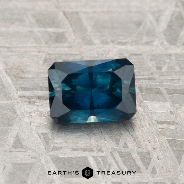 0.96-Carat Montana Sapphire (Heated) A 0.96-carat heated Montana sapphire with faceted edges is displayed on a textured gray surface, with "EARTH'S TREASURY" text at the bottom.