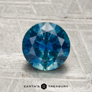 A 1.61-carat heated Montana sapphire with a round brilliant cut rests on a textured gray surface, with "Earth's Treasury" displayed at the bottom.