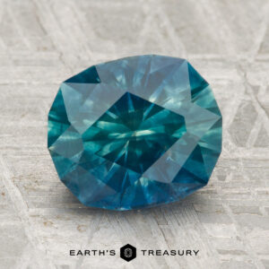 A 3.09-carat heated Montana sapphire with a blue-green hue rests on a textured gray surface, with "EARTH'S TREASURY" at the bottom.