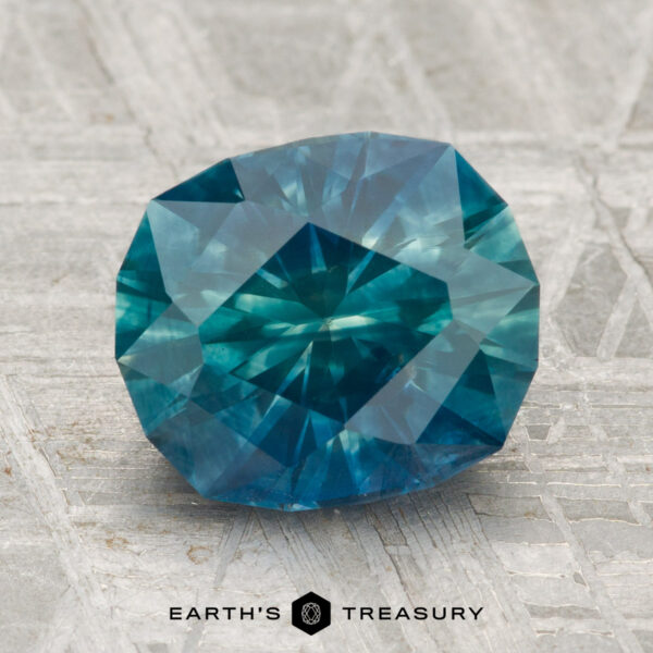 3.09-Carat Montana Sapphire (Heated) A 3.09-carat heated Montana sapphire with a blue-green hue rests on a textured gray surface, with "EARTH'S TREASURY" at the bottom.