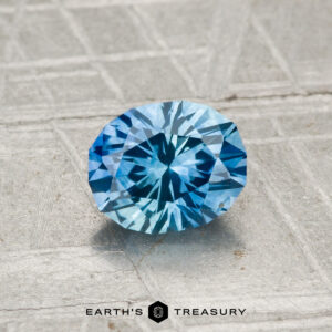 A 1.02-carat heated Montana sapphire with a round brilliant cut is placed on a textured surface, with "EARTH'S TREASURY" displayed at the bottom.