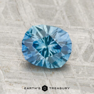 A 1.38-carat heated Montana sapphire with a faceted blue surface rests on gray textured material, with "EARTH'S TREASURY" and a geometric logo displayed below.