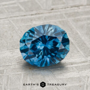 A faceted oval-cut 1.82-carat heated Montana sapphire is displayed on a textured light surface with the text "EARTH'S TREASURY" beneath it.