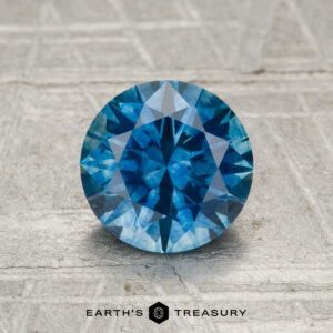 A 1.55-carat heated Montana sapphire, round brilliant-cut and blue, is displayed upright on a textured gray surface with the "Earth's Treasury" logo below it.