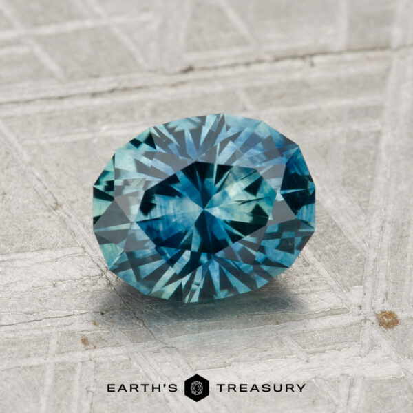 1.78-Carat Montana Sapphire (Heated) A 1.78-carat heated Montana sapphire, round and brilliant-cut, rests on a textured gray surface with "EARTH'S TREASURY" visible at the bottom.