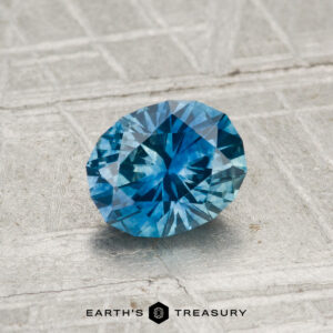 A 1.00-carat heated Montana sapphire rests on a textured gray surface with "Earth's Treasury" and a hexagon logo beneath it.