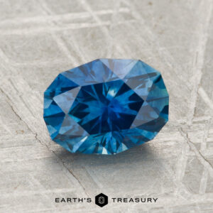 A 2.08-carat heated Montana sapphire with a blue hue rests on a textured gray surface, with the text "EARTH'S TREASURY" at the bottom.