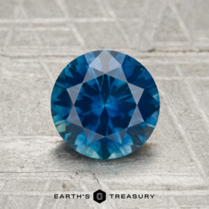 A 1.49-carat faceted Montana sapphire (heated) is displayed on a textured gray surface with "Earth's Treasury" at the bottom.