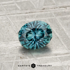 A 1.00-carat heated Montana sapphire with a round brilliant cut is displayed on a textured gray surface, with the "EARTH'S TREASURY" text and logo at the bottom.