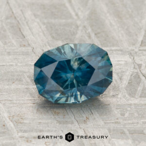 A 1.63-carat heated Montana sapphire with an oval cut and blue-green hue rests on a textured gray surface, with "EARTH'S TREASURY" displayed at the bottom.