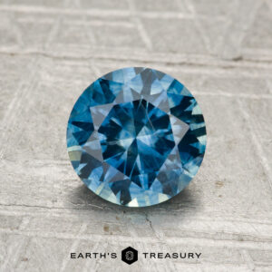 A 1.48-carat round-cut heated Montana sapphire with brilliant facets is displayed on a textured light gray surface, with the Earth’s Treasury logo below the gem.