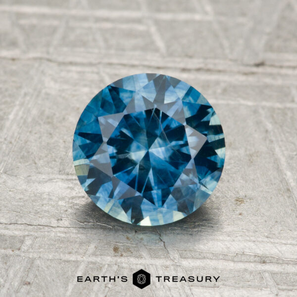 1.48-Carat Montana Sapphire (Heated) A 1.48-carat round-cut heated Montana sapphire with brilliant facets is displayed on a textured light gray surface, with the Earth’s Treasury logo below the gem.