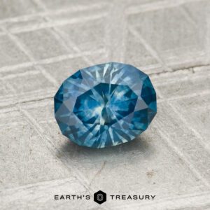 0.99-carat oval-cut heated Montana sapphire with intricate facets shown on a textured light gray surface; "Earth's Treasury" logo appears at the bottom.