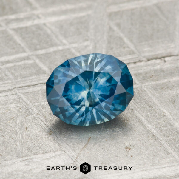 0.99-Carat Montana Sapphire (Heated) 0.99-carat oval-cut heated Montana sapphire with intricate facets shown on a textured light gray surface; "Earth's Treasury" logo appears at the bottom.