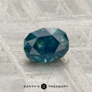 A 1.20-carat heated Montana sapphire, blue-green in color, rests on a textured gray metallic surface with "EARTH'S TREASURY" displayed at the bottom.