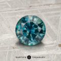 A 1.44-carat heated Montana sapphire, round and blue-green with facets, is displayed on a textured gray surface. "Earth's Treasury" appears at the bottom of the image.