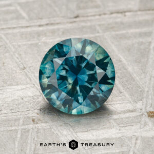 A 1.44-carat heated Montana sapphire, round and blue-green with facets, is displayed on a textured gray surface. "Earth's Treasury" appears at the bottom of the image.