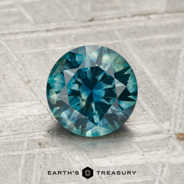 1.44-Carat Montana Sapphire (Heated) A 1.44-carat heated Montana sapphire, round and blue-green with facets, is displayed on a textured gray surface. "Earth's Treasury" appears at the bottom of the image.