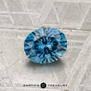 A round, faceted 0.98-carat heated Montana Sapphire with a blue hue rests on a textured gray surface; "EARTH'S TREASURY" logo and text appear at the image's bottom.
