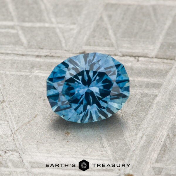0.98-Carat Montana Sapphire (Heated) A round, faceted 0.98-carat heated Montana Sapphire with a blue hue rests on a textured gray surface; "EARTH'S TREASURY" logo and text appear at the image's bottom.