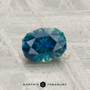 A faceted teal and blue 1.12-carat Montana Sapphire (Heated) rests on a textured gray surface, with "EARTH'S TREASURY" below.