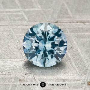 A 1.42-carat heated Montana sapphire with a round, brilliant cut is displayed on a textured silver-gray surface featuring the "Earth's Treasury" logo at the bottom.