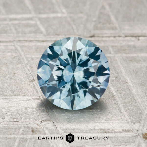1.42-Carat Montana Sapphire (Heated) A 1.42-carat heated Montana sapphire with a round, brilliant cut is displayed on a textured silver-gray surface featuring the "Earth's Treasury" logo at the bottom.