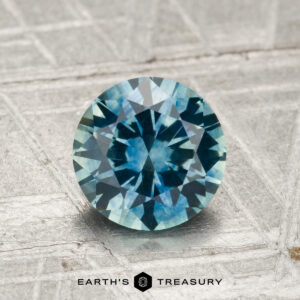 A 1.42-carat heated Montana sapphire with a round brilliant cut and blue-green hue is displayed on a textured silvery surface, featuring the "Earth's Treasury" logo beneath it.