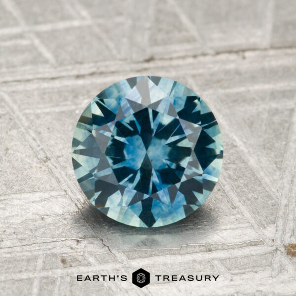 1.42-Carat Montana Sapphire (Heated) A 1.42-carat heated Montana sapphire with a round brilliant cut and blue-green hue is displayed on a textured silvery surface, featuring the "Earth's Treasury" logo beneath it.