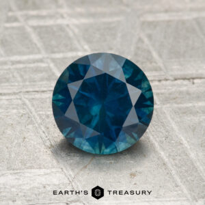 A round, faceted 1.38-carat heated Montana sapphire is displayed on a textured, metallic surface with the text "Earth's Treasury" below.