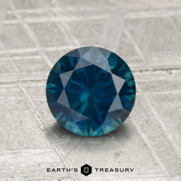 1.38-Carat Montana Sapphire (Heated) A round, faceted 1.38-carat heated Montana sapphire is displayed on a textured, metallic surface with the text "Earth's Treasury" below.
