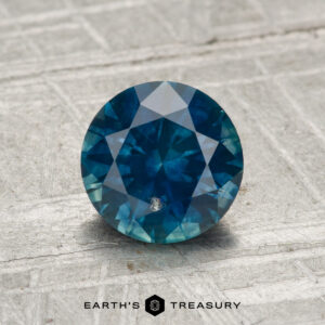 A 1.37-carat heated Montana sapphire, round and blue with faceted cuts, is displayed on a textured gray surface, featuring the text "EARTH'S TREASURY" and a hexagon logo below.