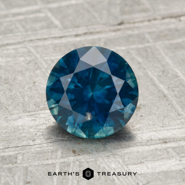 1.37-Carat Montana Sapphire (Heated) A 1.37-carat heated Montana sapphire, round and blue with faceted cuts, is displayed on a textured gray surface, featuring the text "EARTH'S TREASURY" and a hexagon logo below.