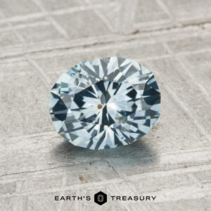 A faceted oval light blue 1.42-carat heated Montana sapphire rests on a textured gray surface with "EARTH'S TREASURY" at the bottom.