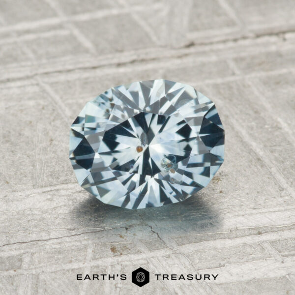 1.42-Carat Montana Sapphire (Heated) A faceted oval light blue 1.42-carat heated Montana sapphire rests on a textured gray surface with "EARTH'S TREASURY" at the bottom.