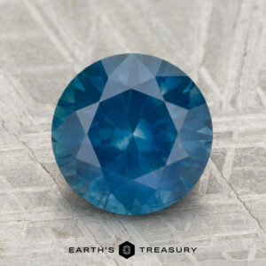 A 3.23-carat heated round brilliant-cut Montana sapphire is displayed on a textured gray surface, featuring the "Earth's Treasury" logo at the bottom.