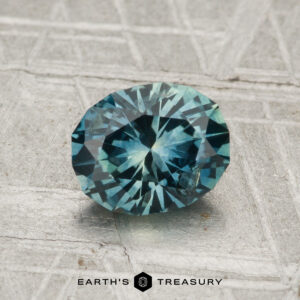 A 1.40-carat heated Montana sapphire with blue-green facets rests on a textured gray surface, "EARTH'S TREASURY" printed below.