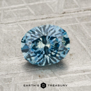A 1.39-carat heated Montana sapphire, round brilliant-cut and blue, is shown on a textured gray surface with the "Earth's Treasury" logo at the bottom.